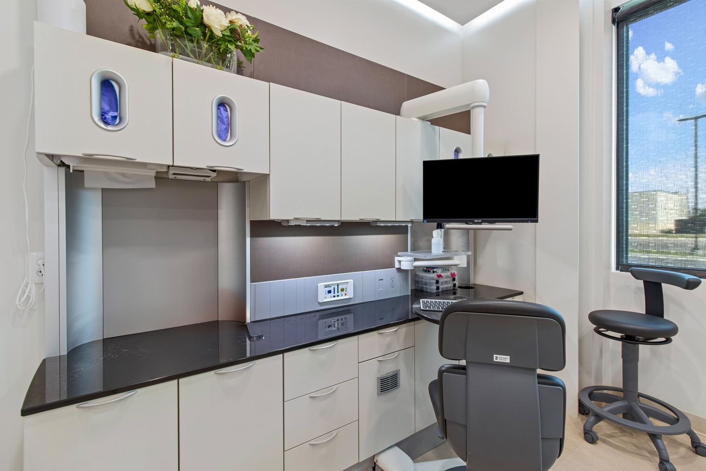 Operatory cabinetry in cream and taupe with a black worktop, a wall monitor and a window beside the chair