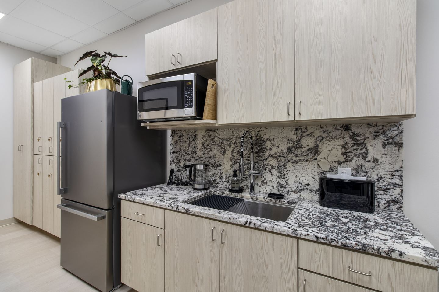 Staff kitchen with pale oak cabinets, a marbled granite splashback and a stainless refrigerator