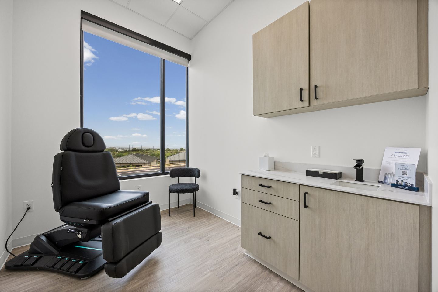 Treatment room with a black powered treatment chair, oak cabinetry and a tall window