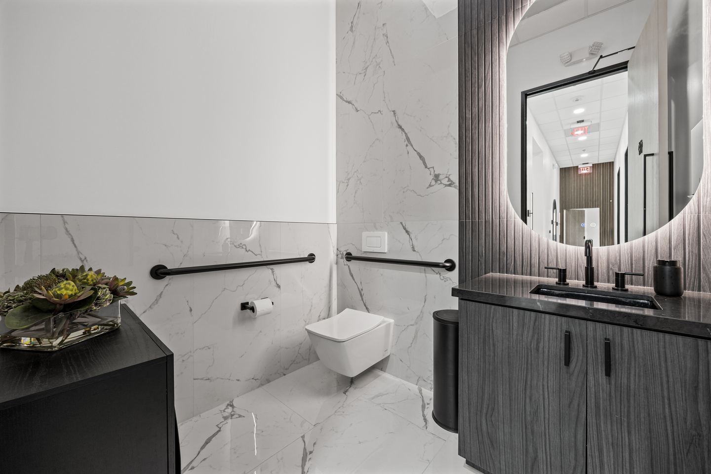 Washroom in white marble tile with a backlit round mirror and a dark wood vanity